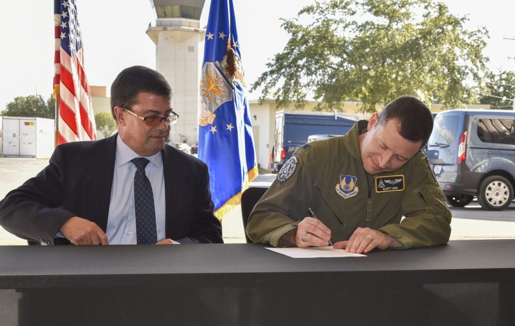 A man in a suit and a man in an air force jumpsuit sit behind a table signing a document